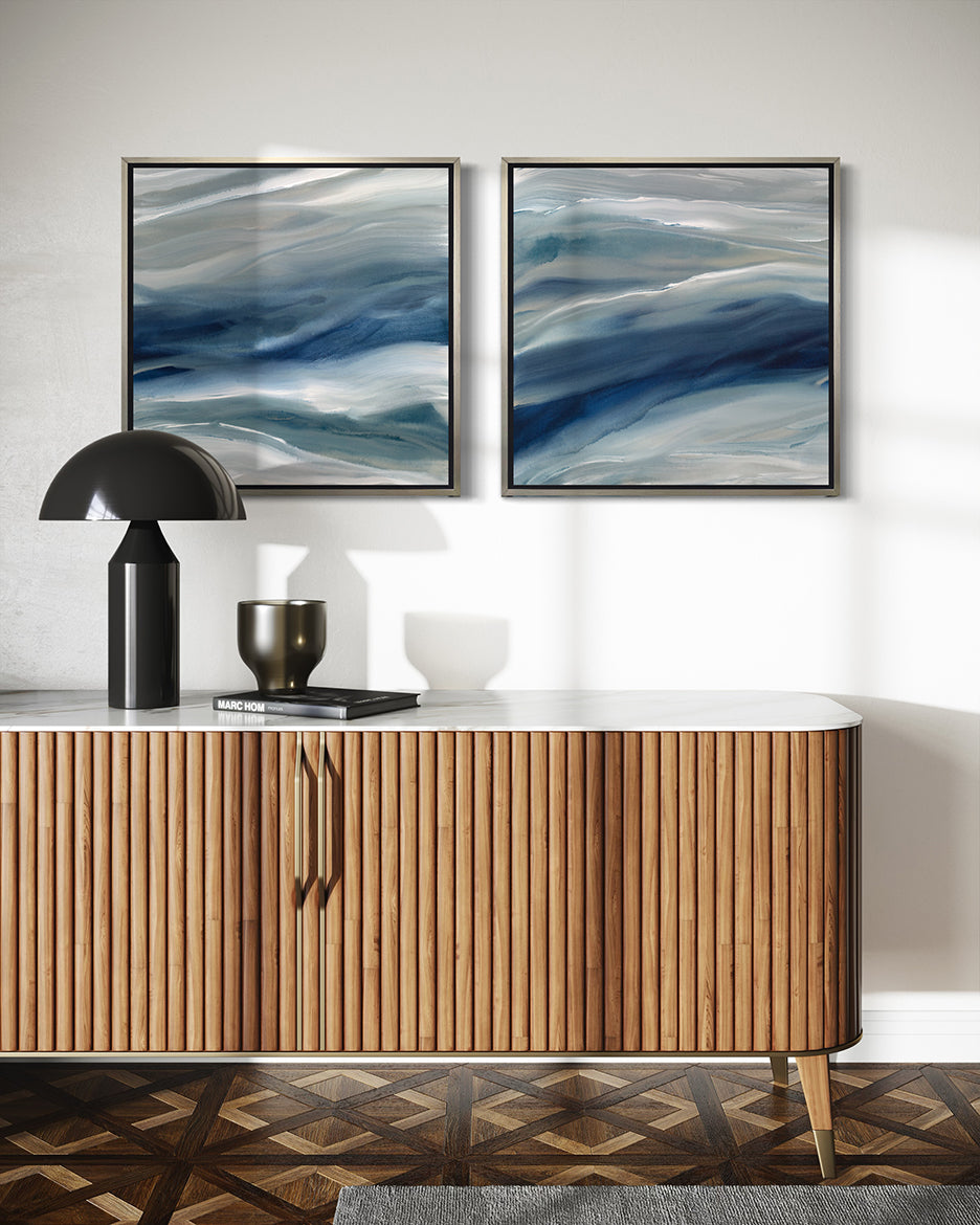 Two blue, teal, beige and white abstract prints in silver floater frames hang on a white wall above a midcentury modern TV cabinet with a black lamp, vase and book.