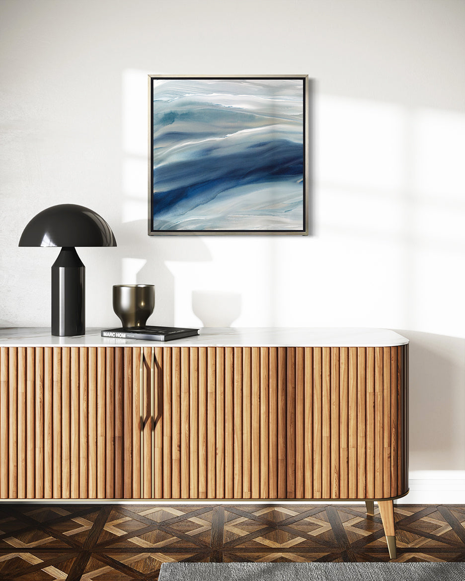 A blue, teal, beige and white abstract prints in silver floater frames hang on a white wall above a midcentury modern TV cabinet with a black lamp, vase and book.