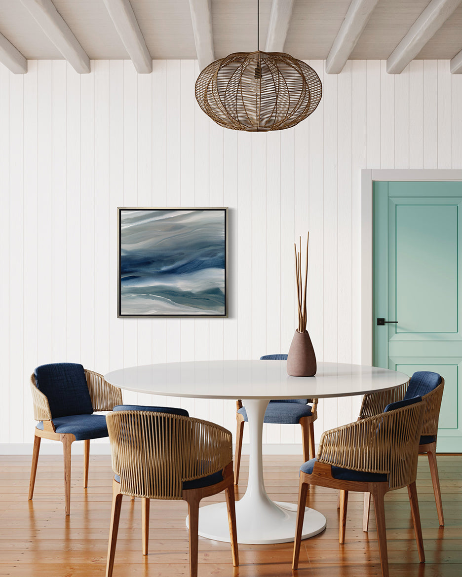 A blue, teal, beige and white abstract print in a silver floater frame hangs on a white wall in a kitchen with a white pedestal table and blue chairs.