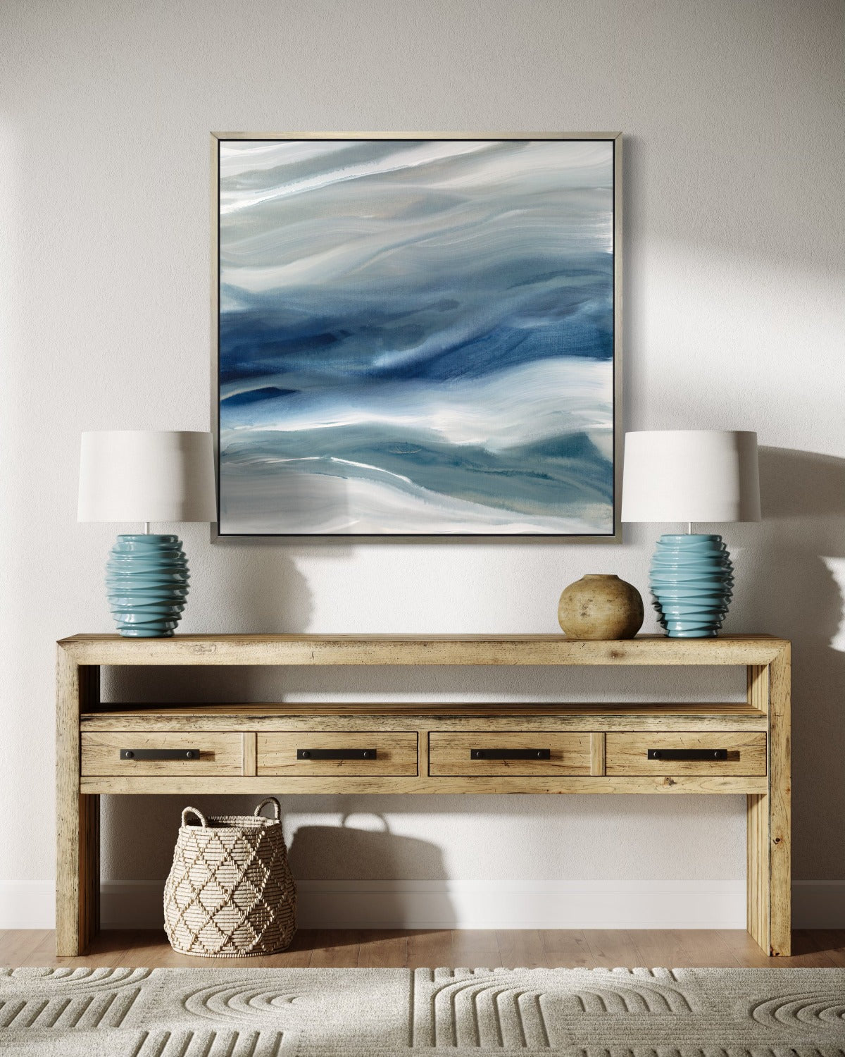 A blue, teal, beige and white abstract print in a silver floater frame hangs on a white wall above a brown wooden credenza with 2 teal lamps and ceramic pot. A wicker basket sits on the floor to the left under the table.