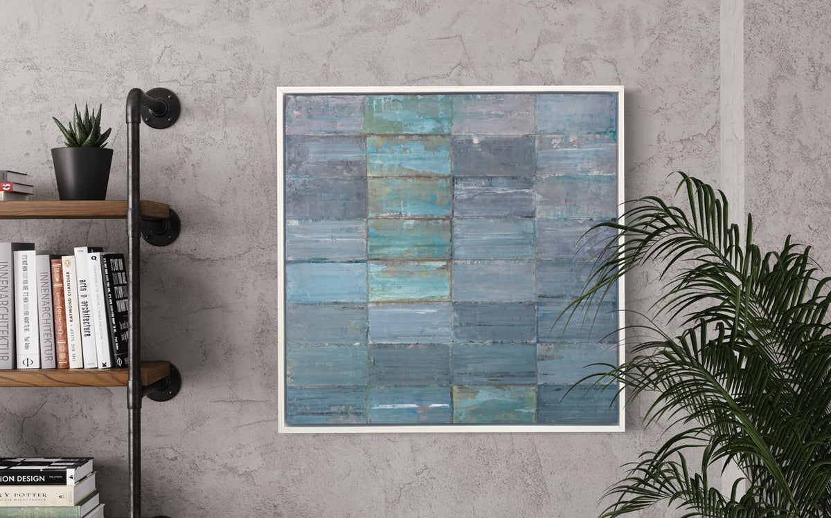 An abstract geometric slate blue and sea foam green limited edition art print by Ned Martin framed in a white frame hangs on a grey wall between a potted plant and some bookshelves