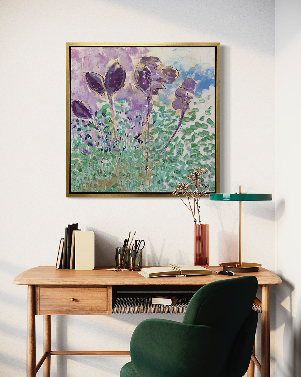 A purple, green and blue abstract floral print in a gold floater frame hangs on a white wall above a natural wood desk with books, a modern lamp, a cylindrical vase and a green armchair.