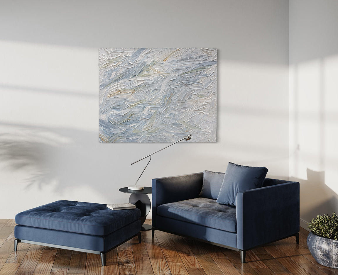 A blue, grey, white, stone, teal, and yellow ochre thickly textured abstract painting by Teodora Guererra on a white wall in natural light hangs over a blue oversized chair with ottoman. A contemporary end table is to the side of the chair with a modern light. Also, a plant in a grey pot sits on the floor to the right of the blue chair.
