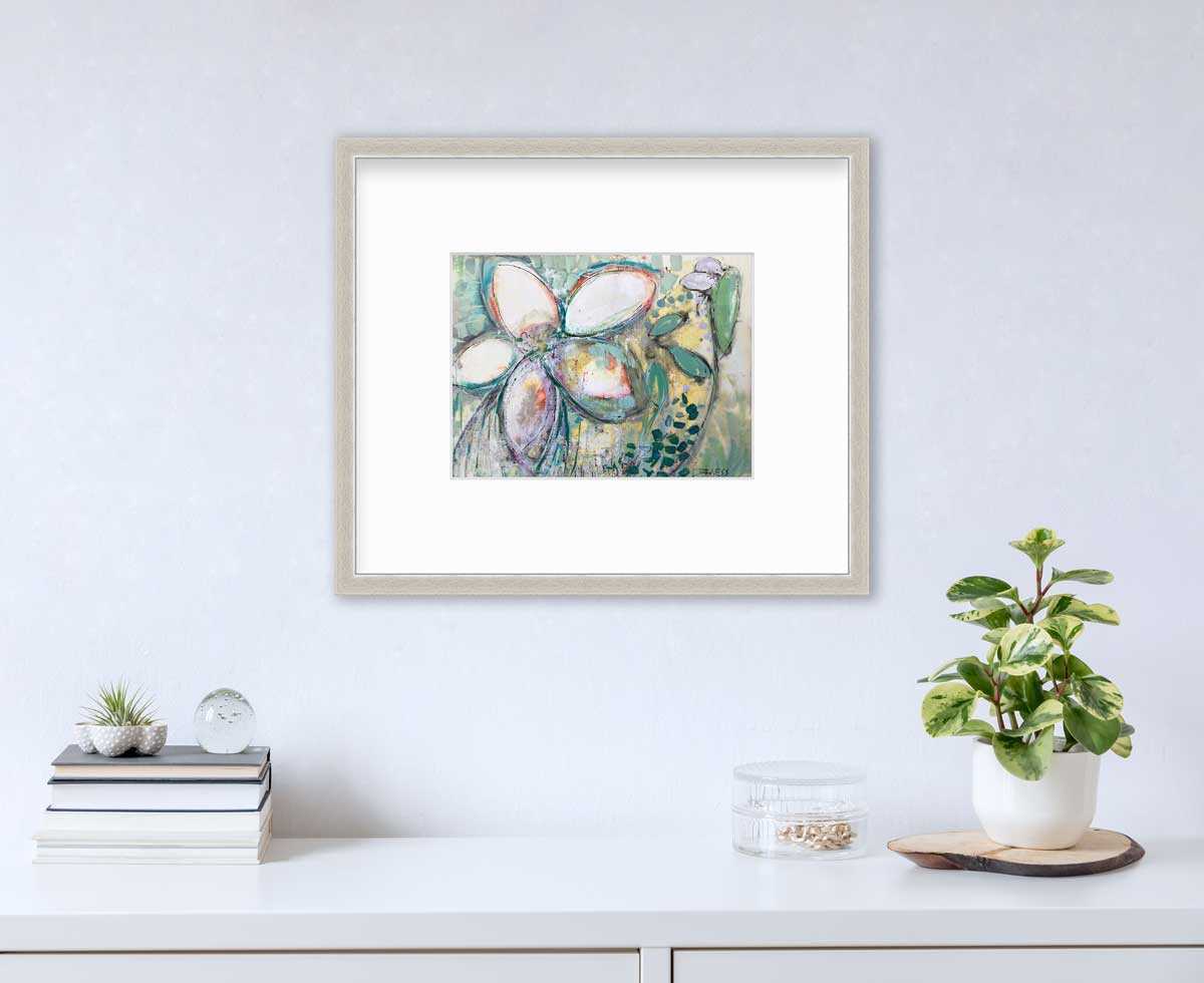 An abstract floral print in a silver frame with a mat hangs on a white wall above a white surface with a stack of books, a clear container with gold accessories and a potted plant.