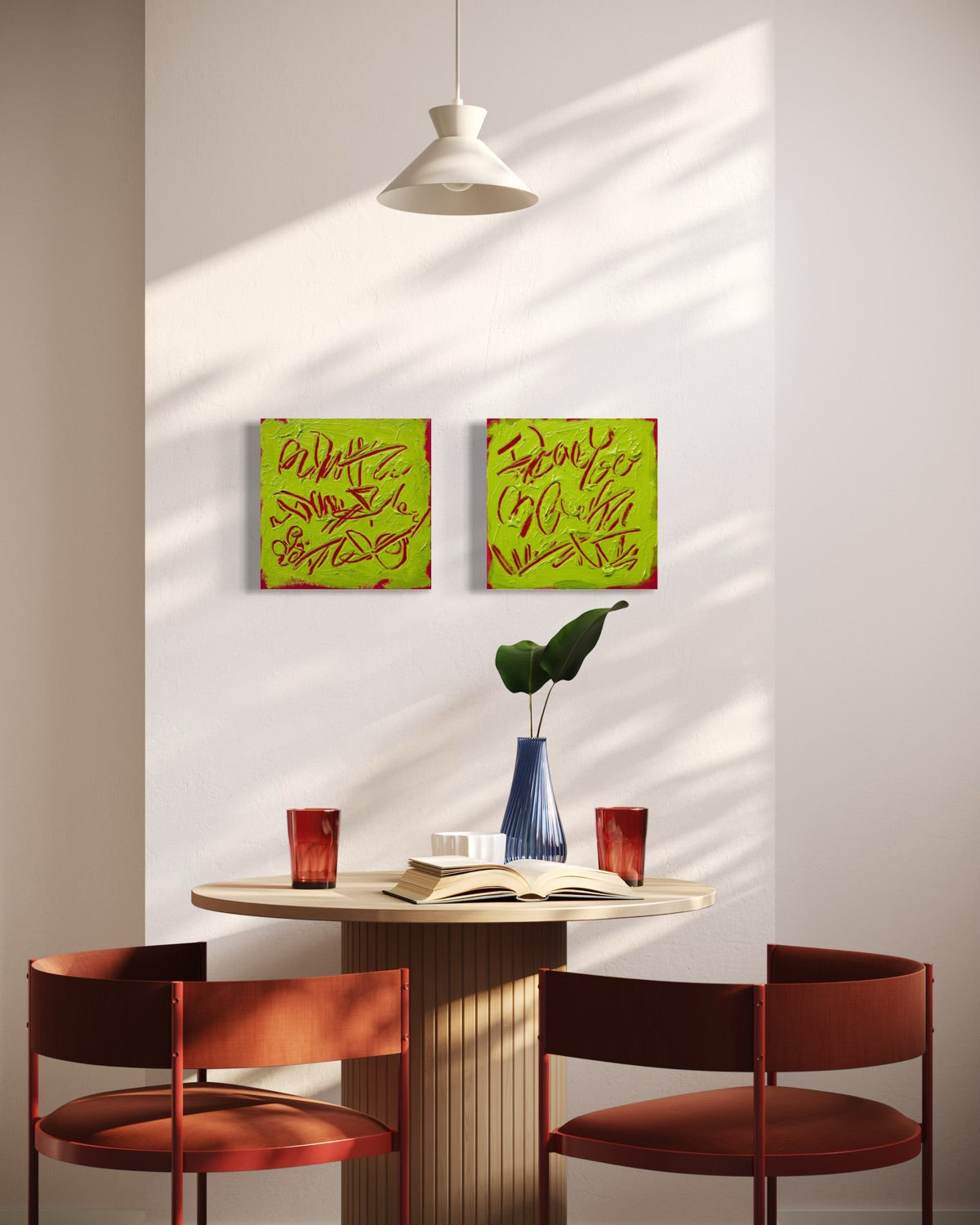 Two square abstract text-based paintings with textured chartreuse over hot raspberry pink graffiti-style lettering, hang on a light gray wall above a round dining table with two rust-red chairs. A blue vase with a green leaf and an open book sit on the table, with sunlight casting shadows across the wall.