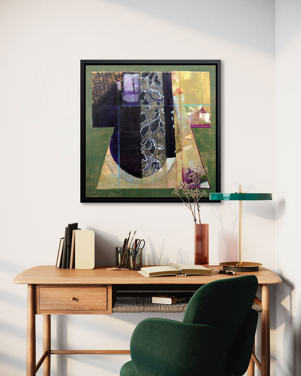 A green, gold and black abstract print in a black floater frame hangs on a white wall above a natural wood desk with accessories and a deep green chair.