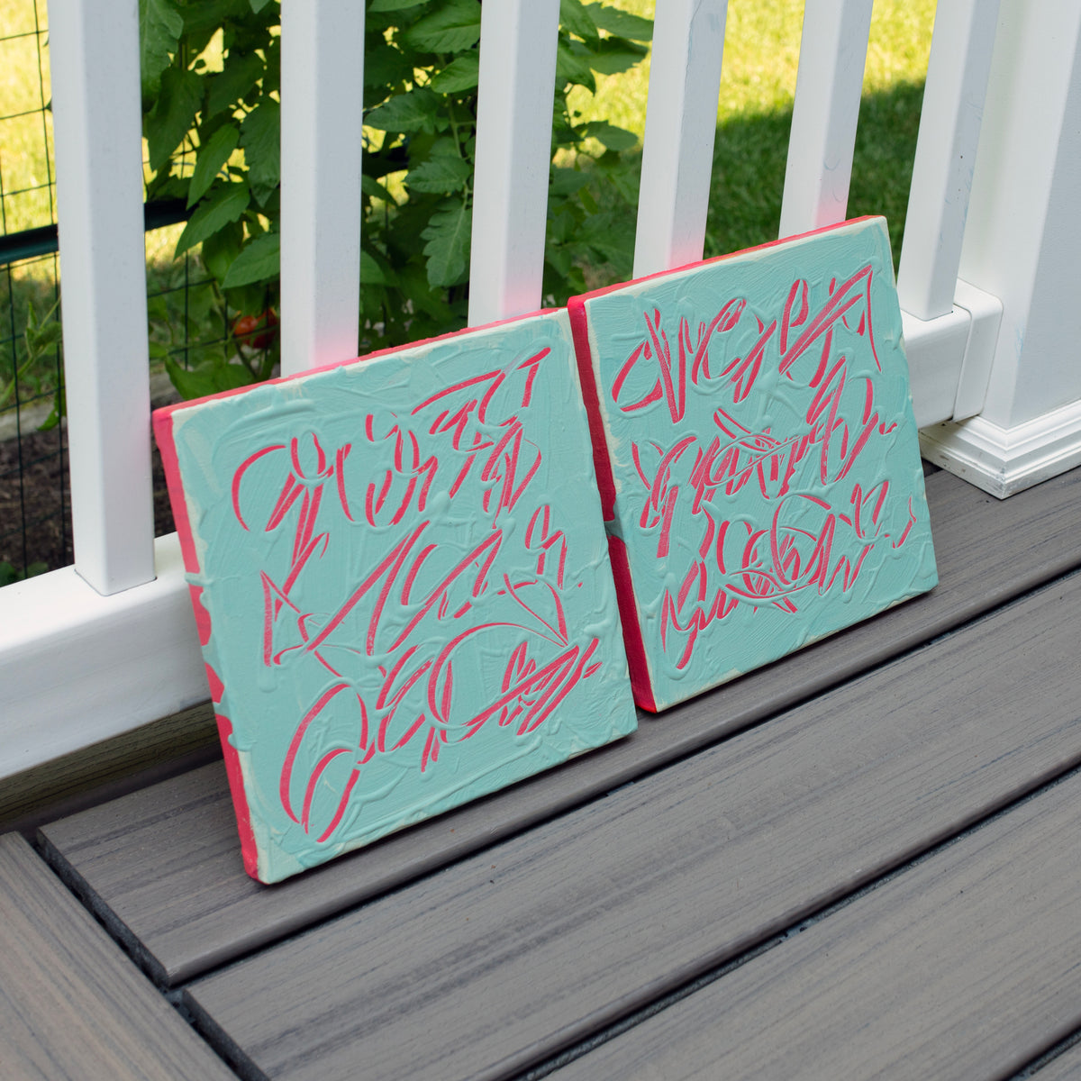 Two abstract text based paintings displayed side-by-side on a gray wooden deck in front of a white fence with green foliage in the background.