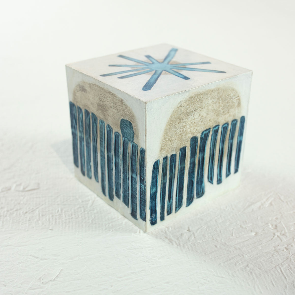A white cube painted with blue and beige geometric shapes rests on a white surface.