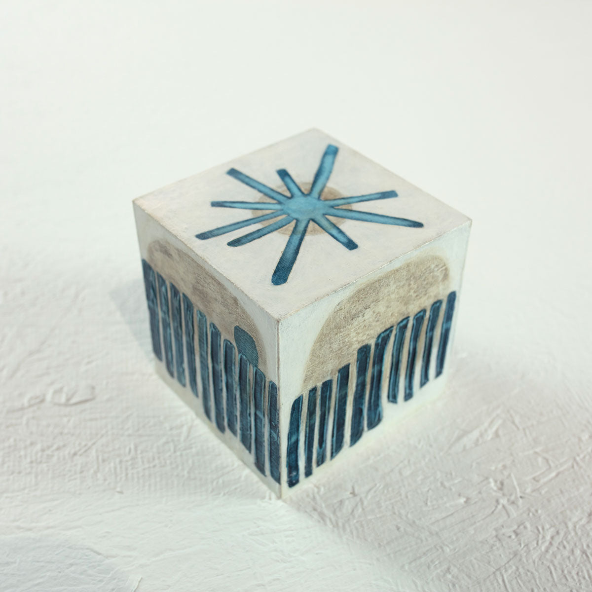 A white cube painted with blue and beige geometric shapes rests on a white surface.