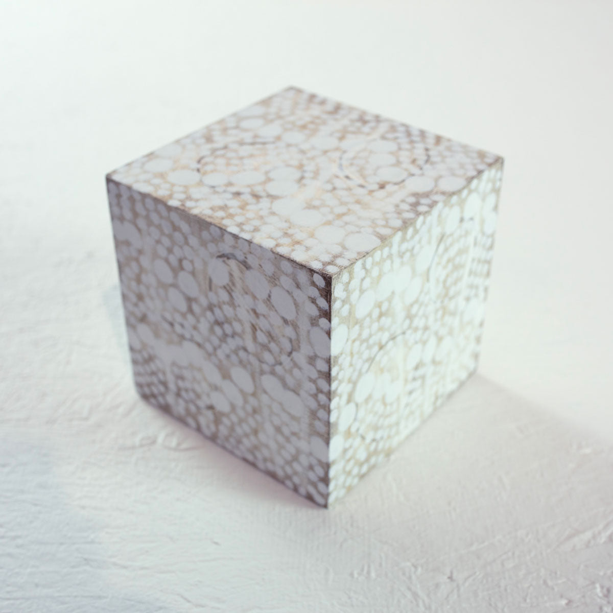 A biege wooden cube painted with small white circles rests on a white surface.