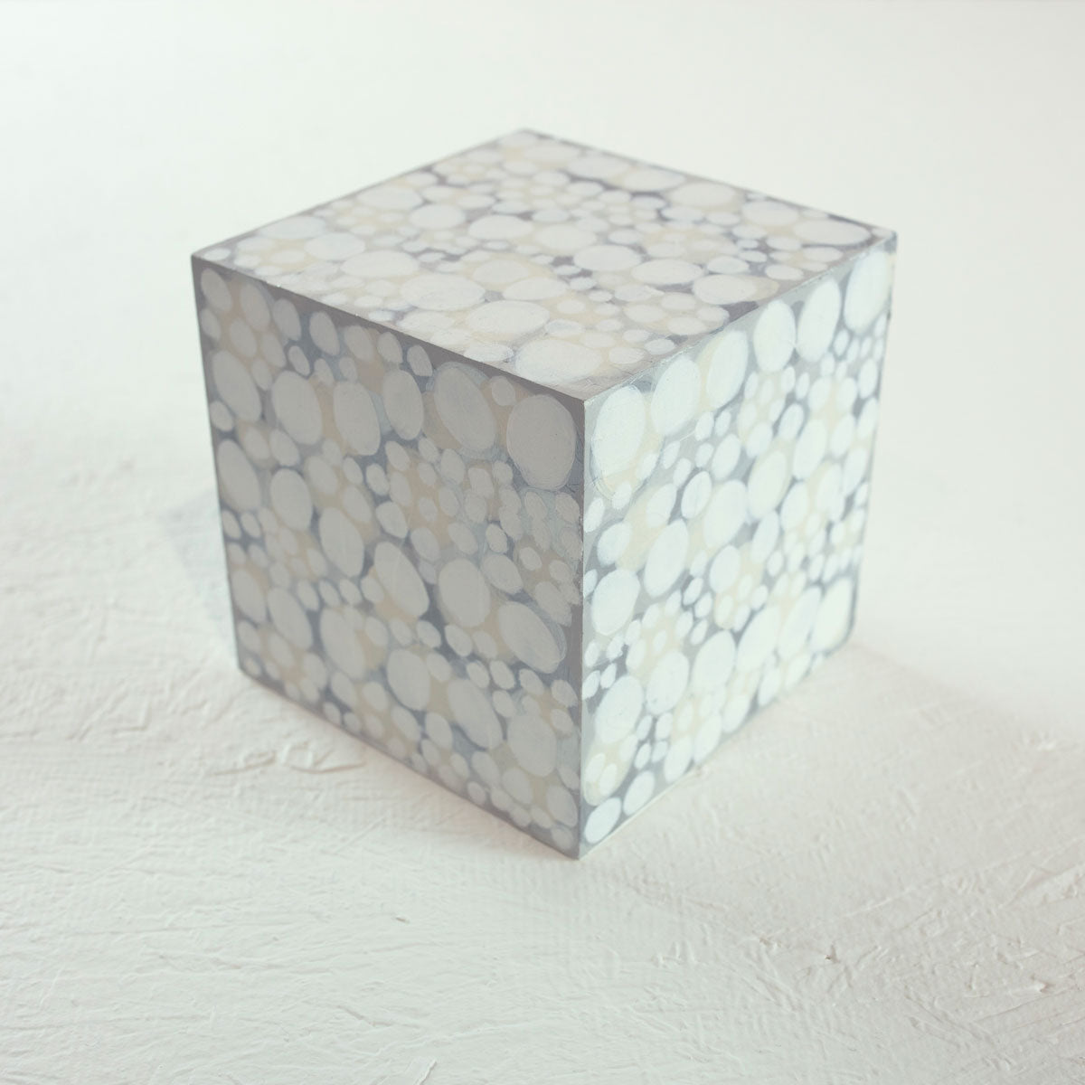 A wooden cube painted with overlapping white, beige, and grey circles rests on a white surface.