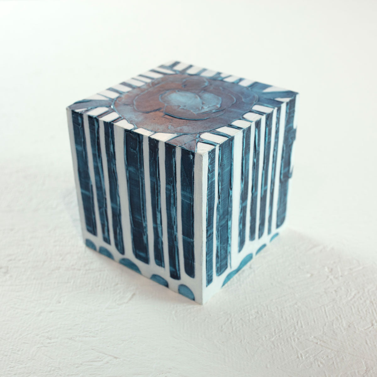 A white cube with a blue circle on the top and textured, geometric rectangles that extend over the sides of the cube rests on a white surface.