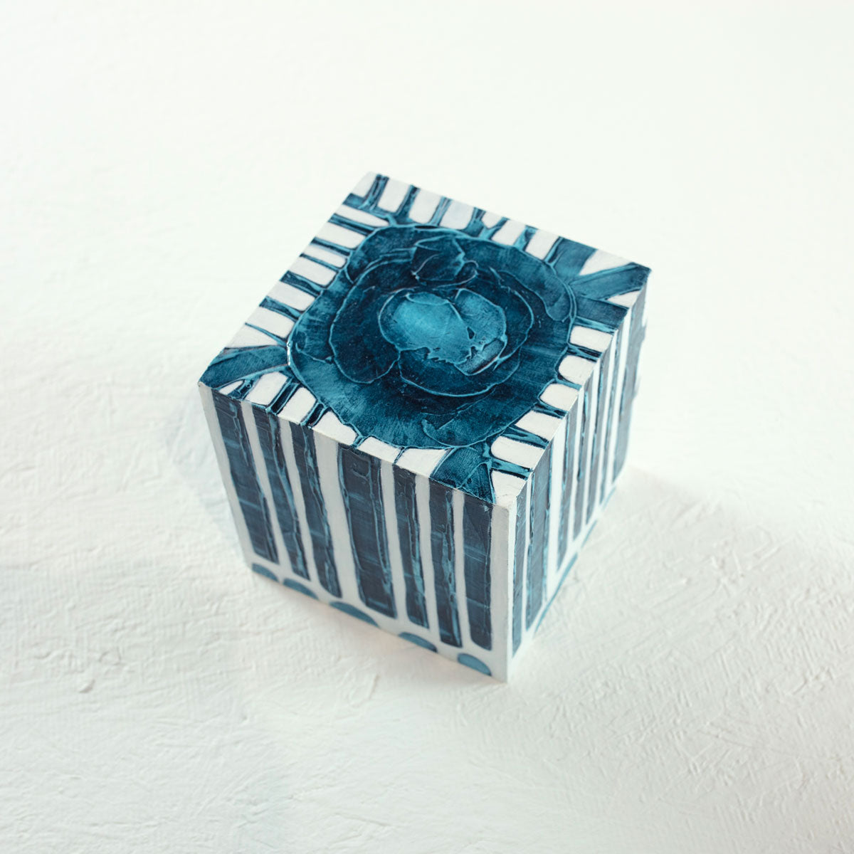 A white cube with a blue circle on the top and textured, geometric rectangles that extend over the sides of the cube rests on a white surface.