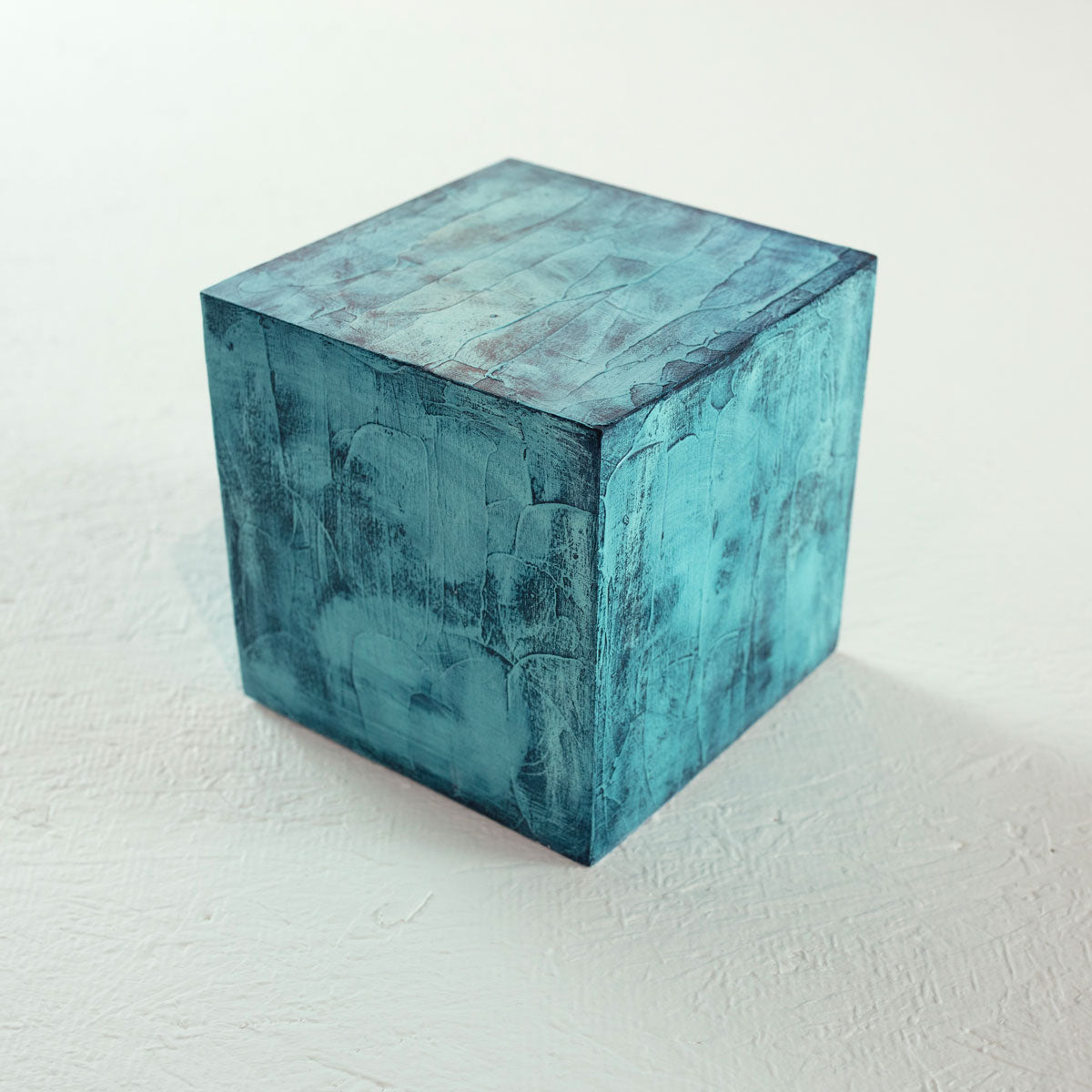 A painted textured turquoise cube rests on a white surface.