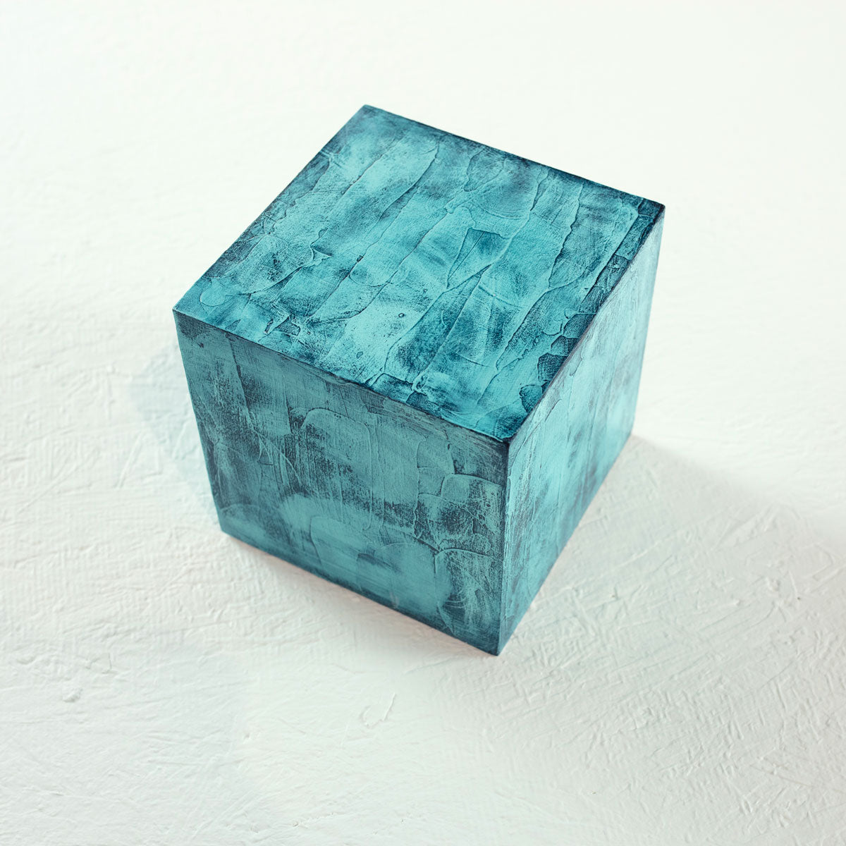 A painted textured turquoise cube rests on a white surface.