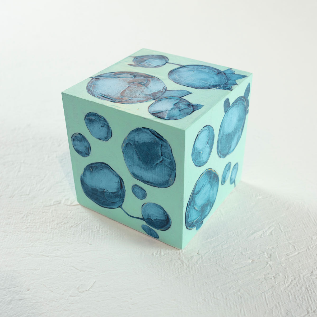 A cube painted turquoise with imperfect blue circles rests on a white surface.