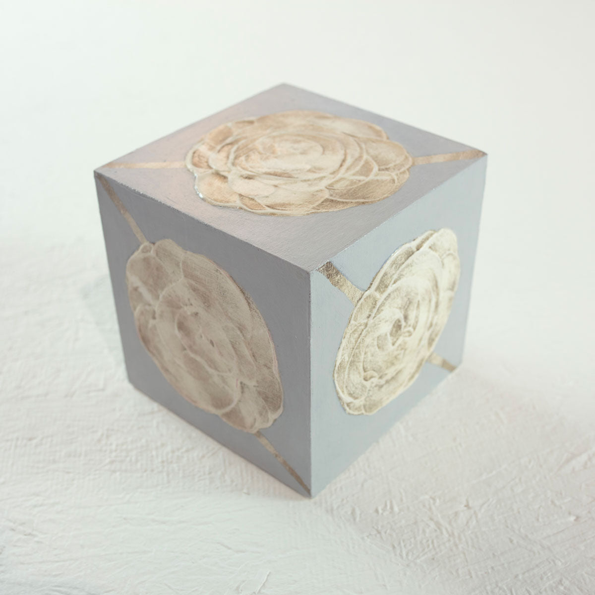 A grey cube with golden beige floral shapes painted on each face of the cube rests on a white surface.