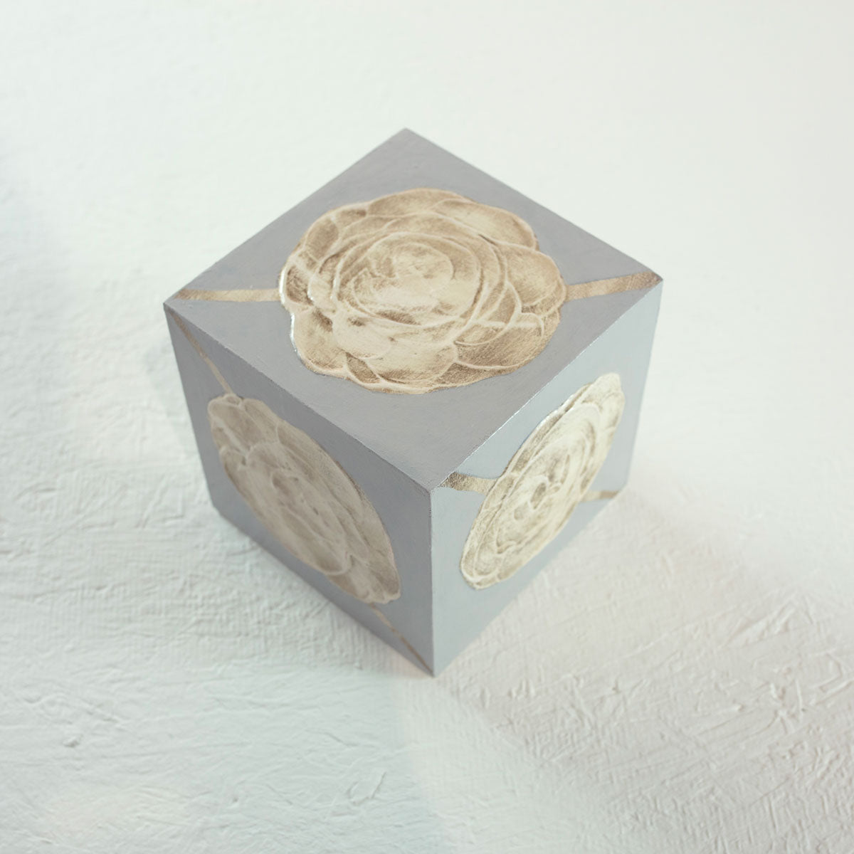 A grey cube with golden beige floral shapes painted on each face of the cube rests on a white surface.