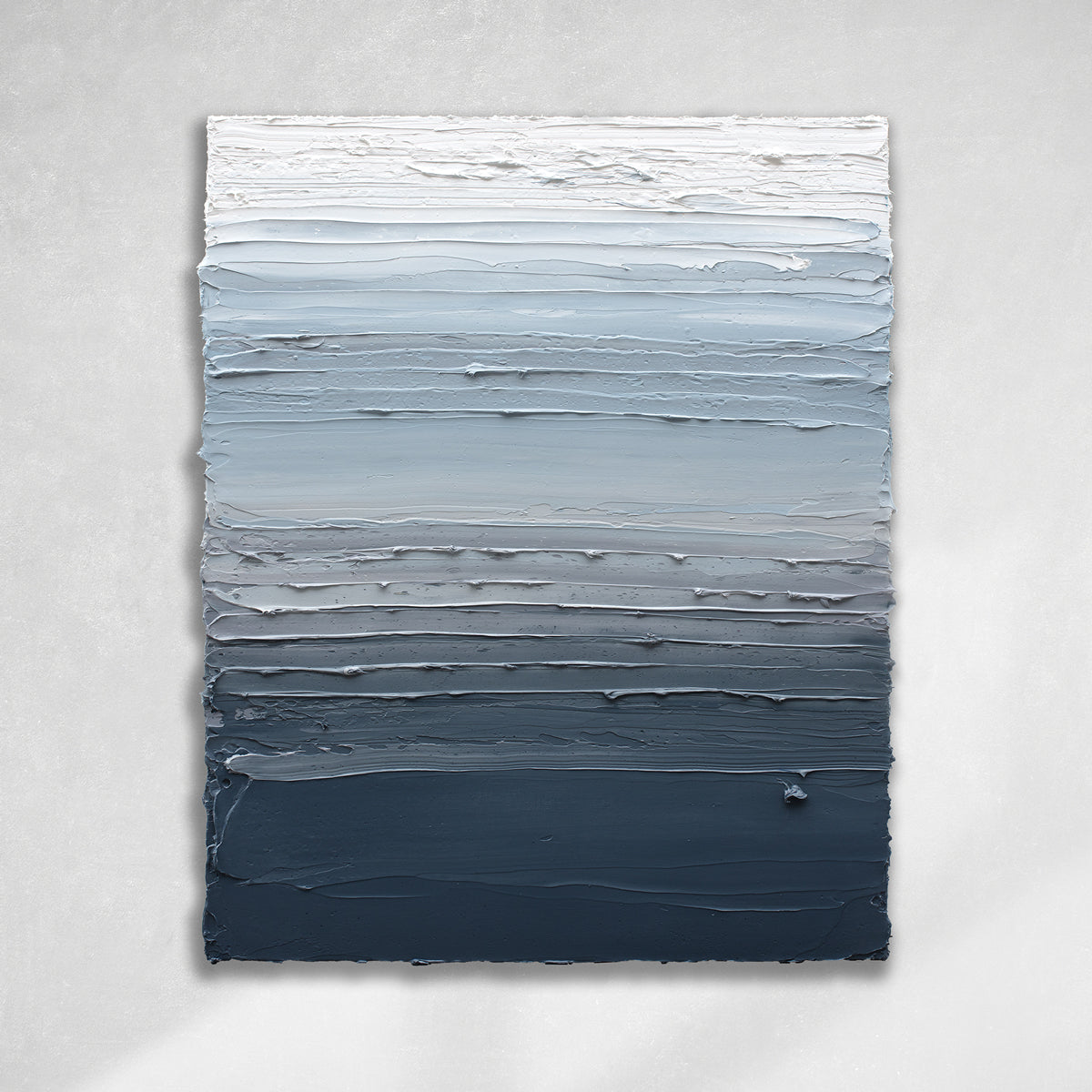 A vertical thickly textured abstract painting in bands of dark blue grey, light blue, blue green, and white hanging on a white wall in natural light by artist Teodora Guererra.