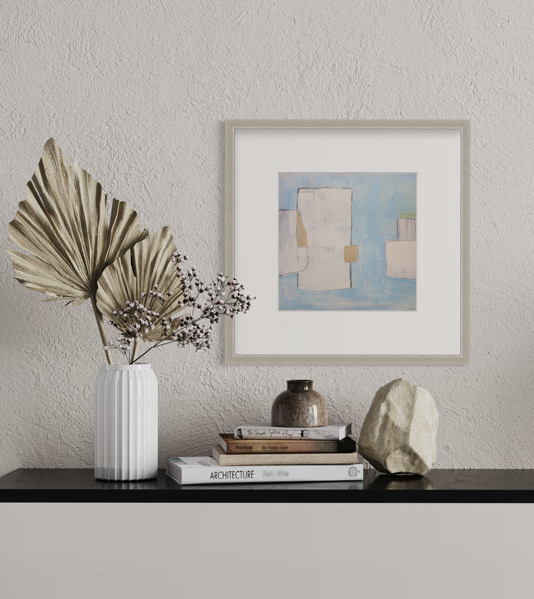 A blue, white and beige abstract print in a silver frame with a mat hangs on a white wall above a black surface with a stack of books, a rock paperweight and a white vase with decorative stems.