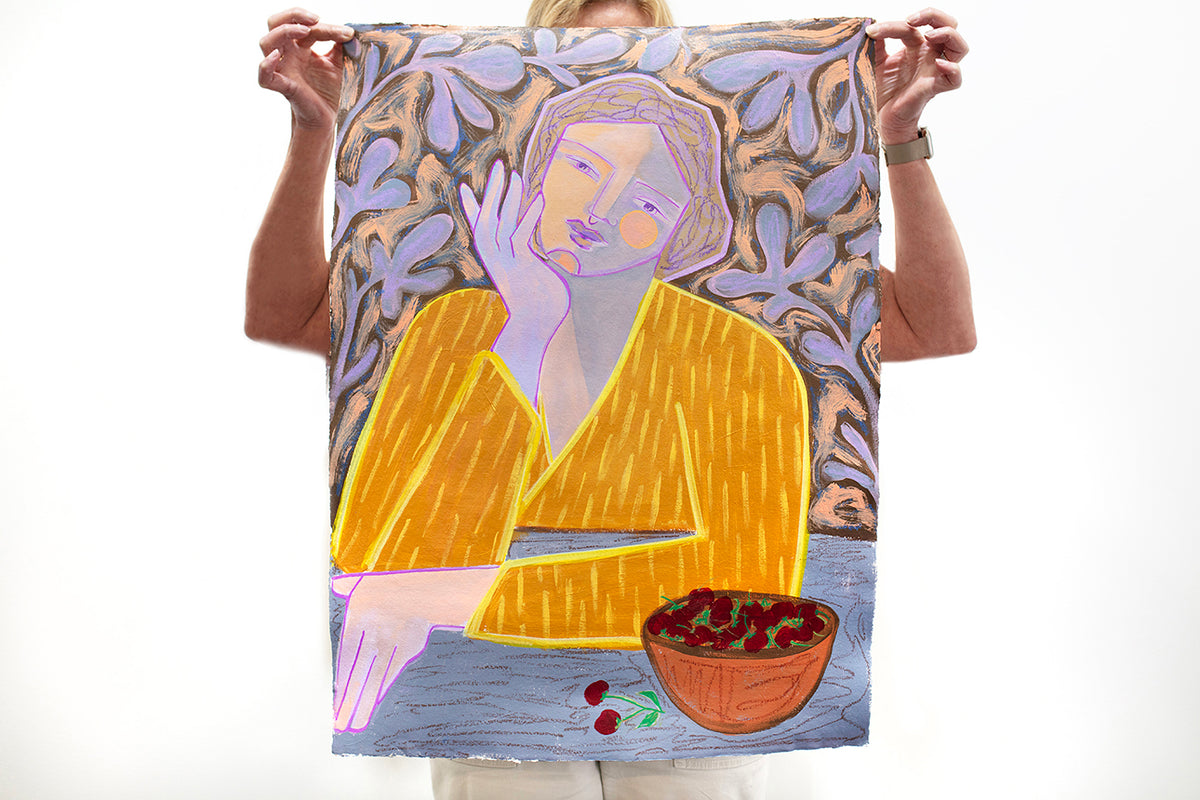 A woman holds a colorful abstract painting of a woman wearing a yellow shirt on paper in front of a white wall.