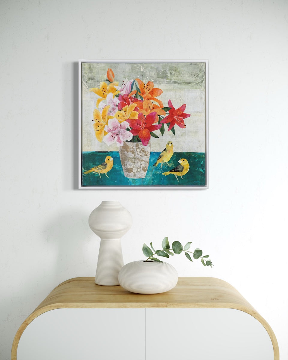 A framed square collage painting depicting a colorful bouquet of flowers in a vase with yellow birds, displayed above a light wood console table with white ceramic vases against a white wall.