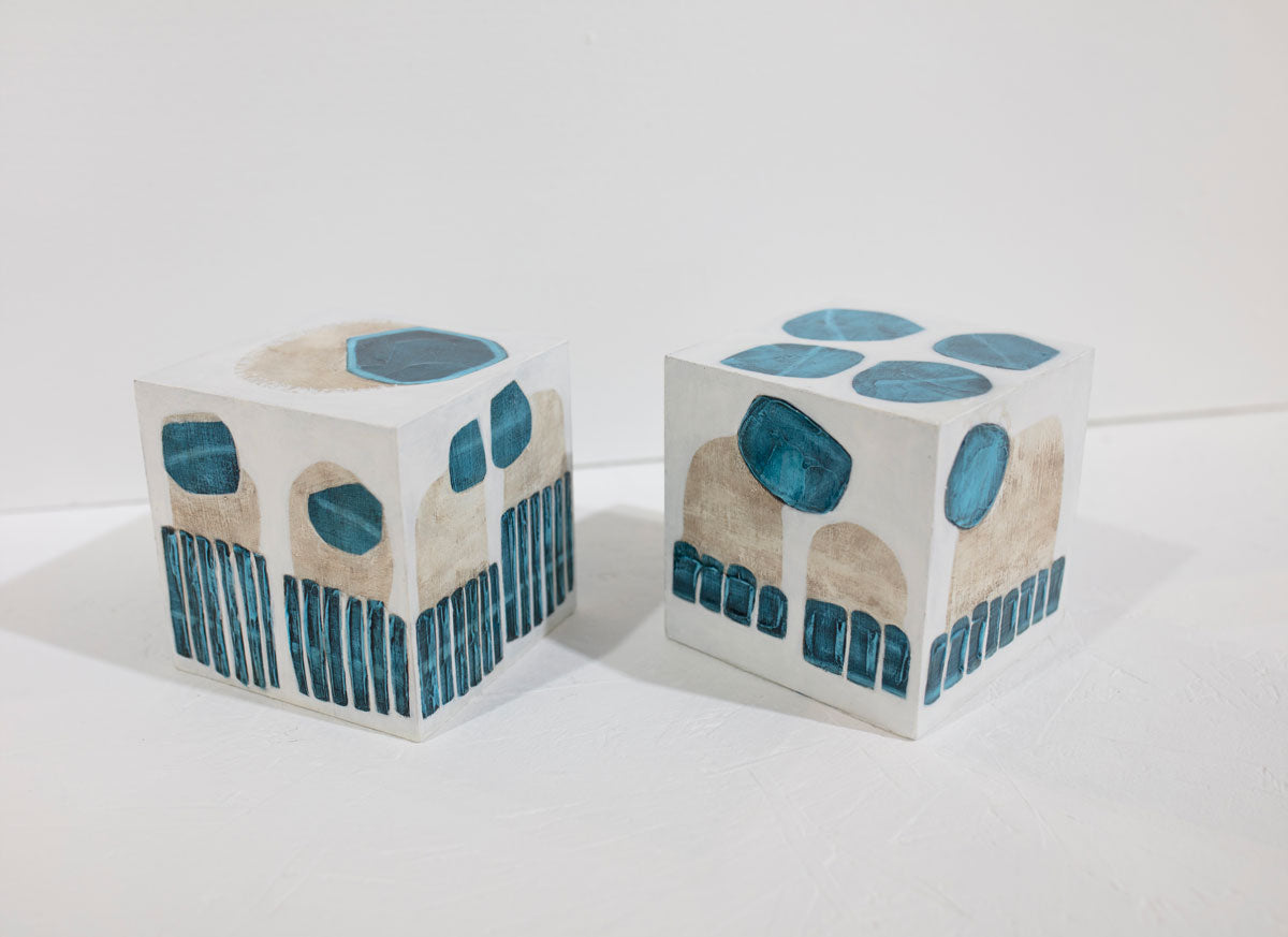 Two white cubes painted with blue and beige shapes sit on a white surface in front of a white wall.