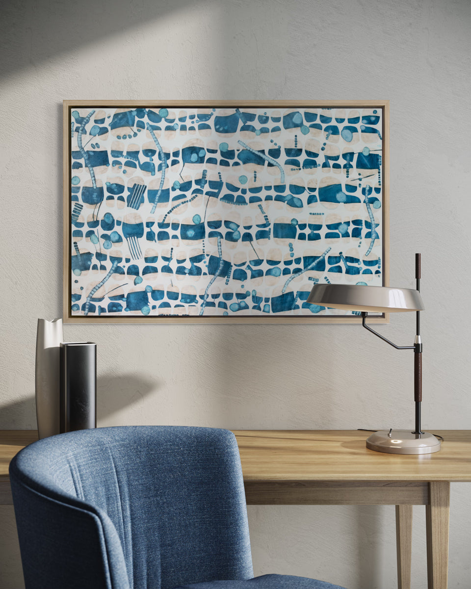 A blue, teal and beige geometric abstract print in a white oak wood floater frame hangs on a white wall above a natural wood desk with a small lamp, two vase and a blue cushioned chair.