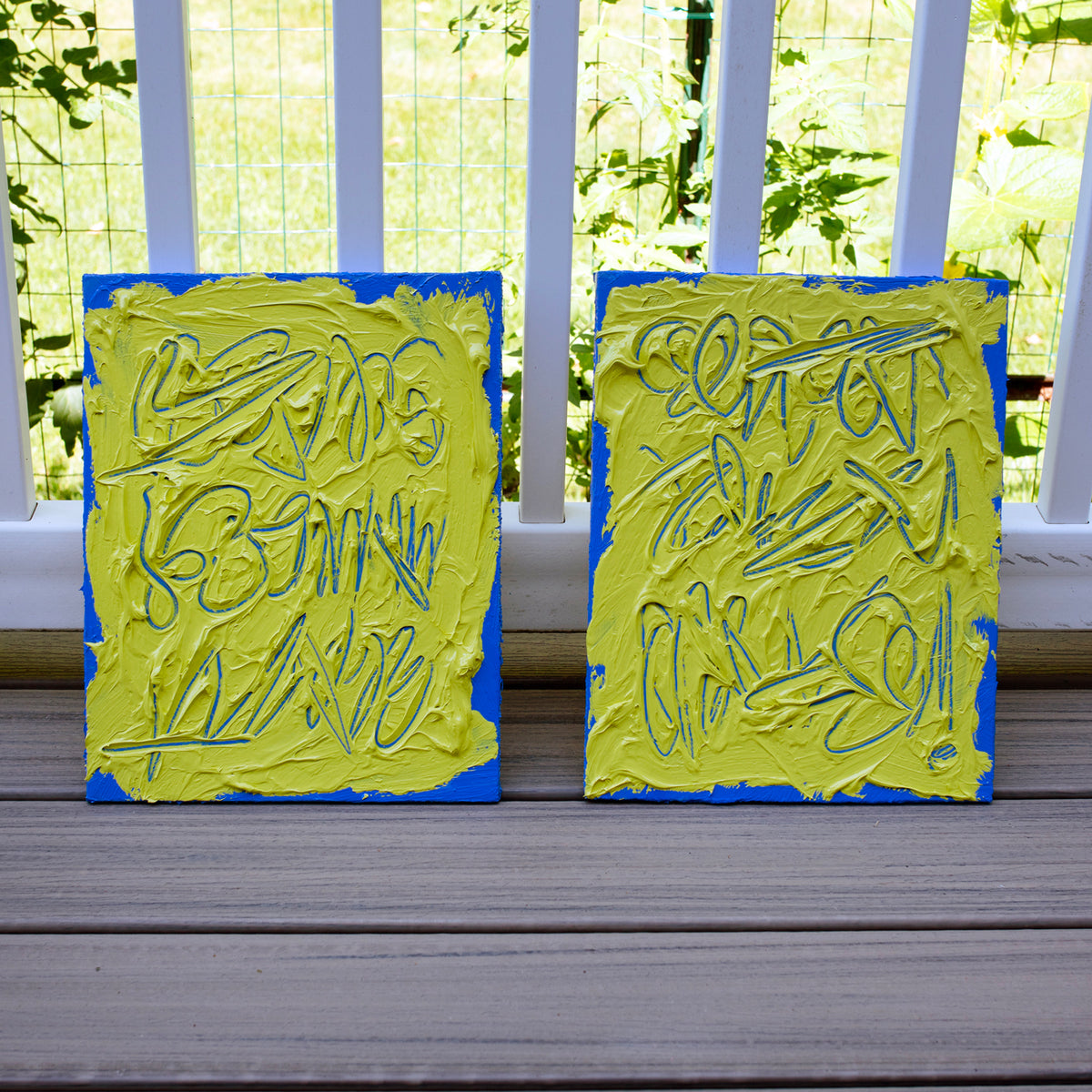 Two abstract text based painting featuring a thick, yellow-green impasto texture with blue abstract scribbled lines and a bright blue background. They are displayed on a wooden deck in front of a white railing with green foliage behind.