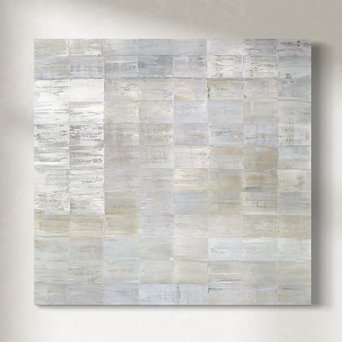 Abstract painting with a grid of rectangular panels in various shades of white, gray and gold hanging on a light gray wall.