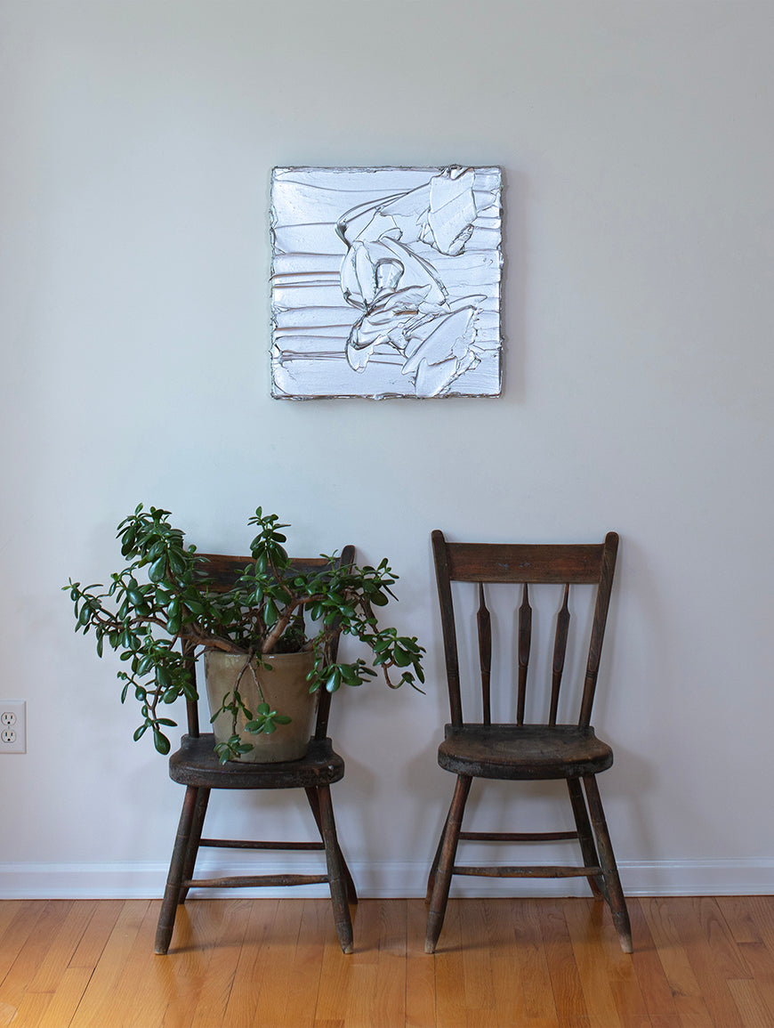 A silver metallic textured painting by Teodora Guererra hanging on a wall in the artists studio above 2 wooden chairs, one with a yucca plant.