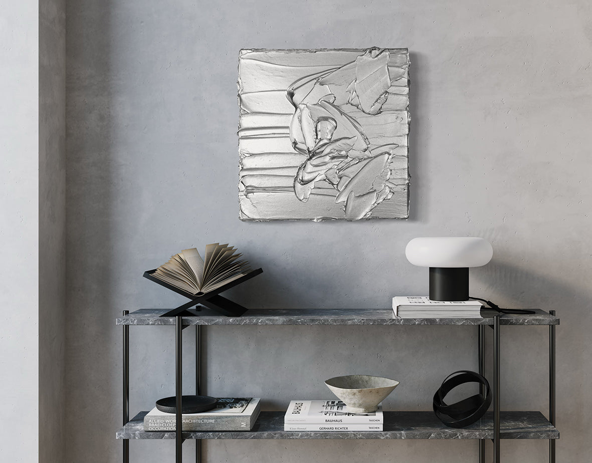 A silver metallic textured painting by Teodora Guererra hanging on a wall above a marble shelf unit with books and decorative objects.