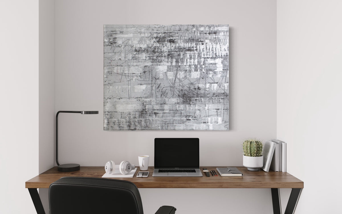 A black and silver abstract painting hangs on a white wall above a minimalist wood desk with lap top, book, and table lamp.