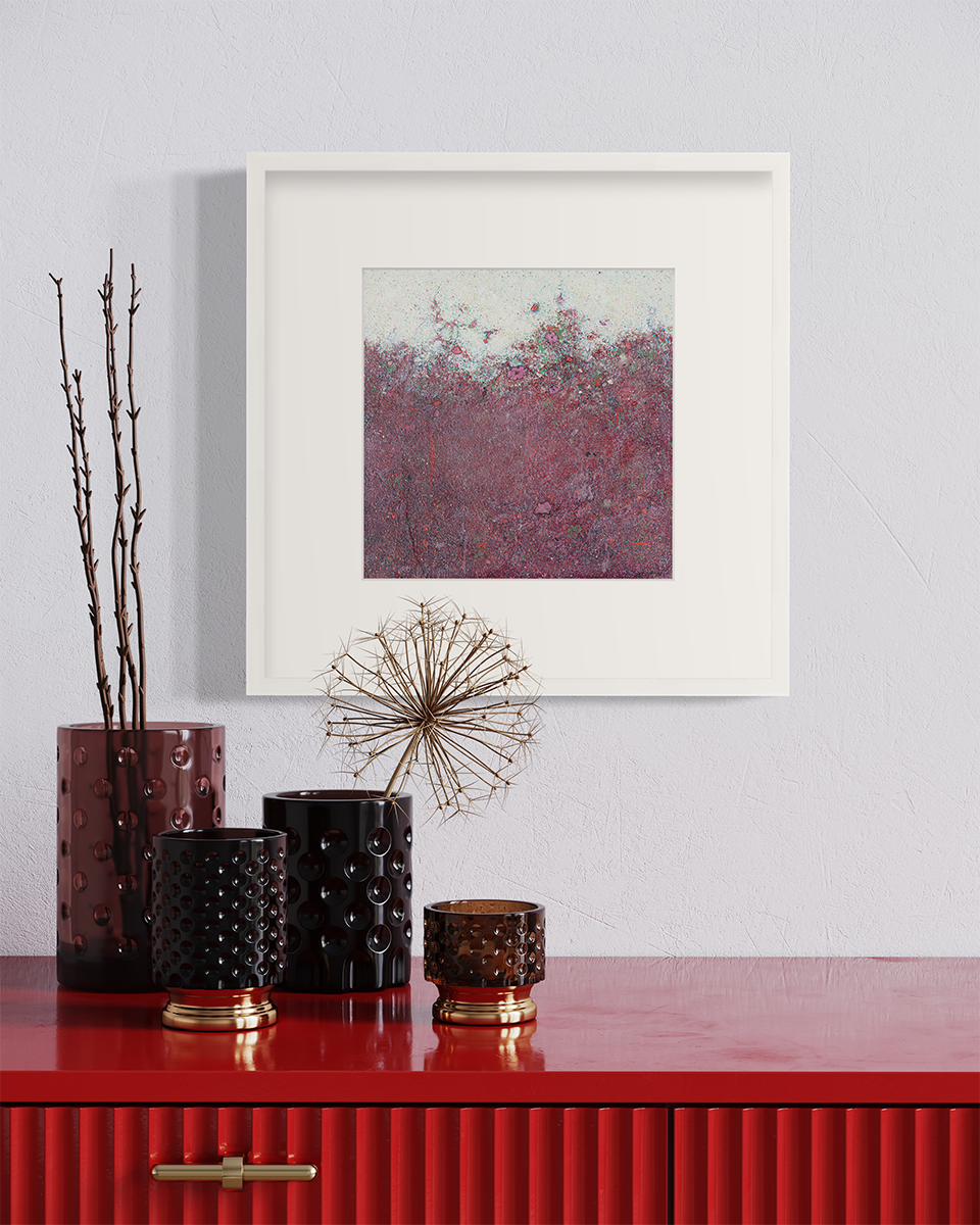A dark red abstract landscape print in a white frame with a mat hangs on a white wall above a red surface with various vases with decorative stems.