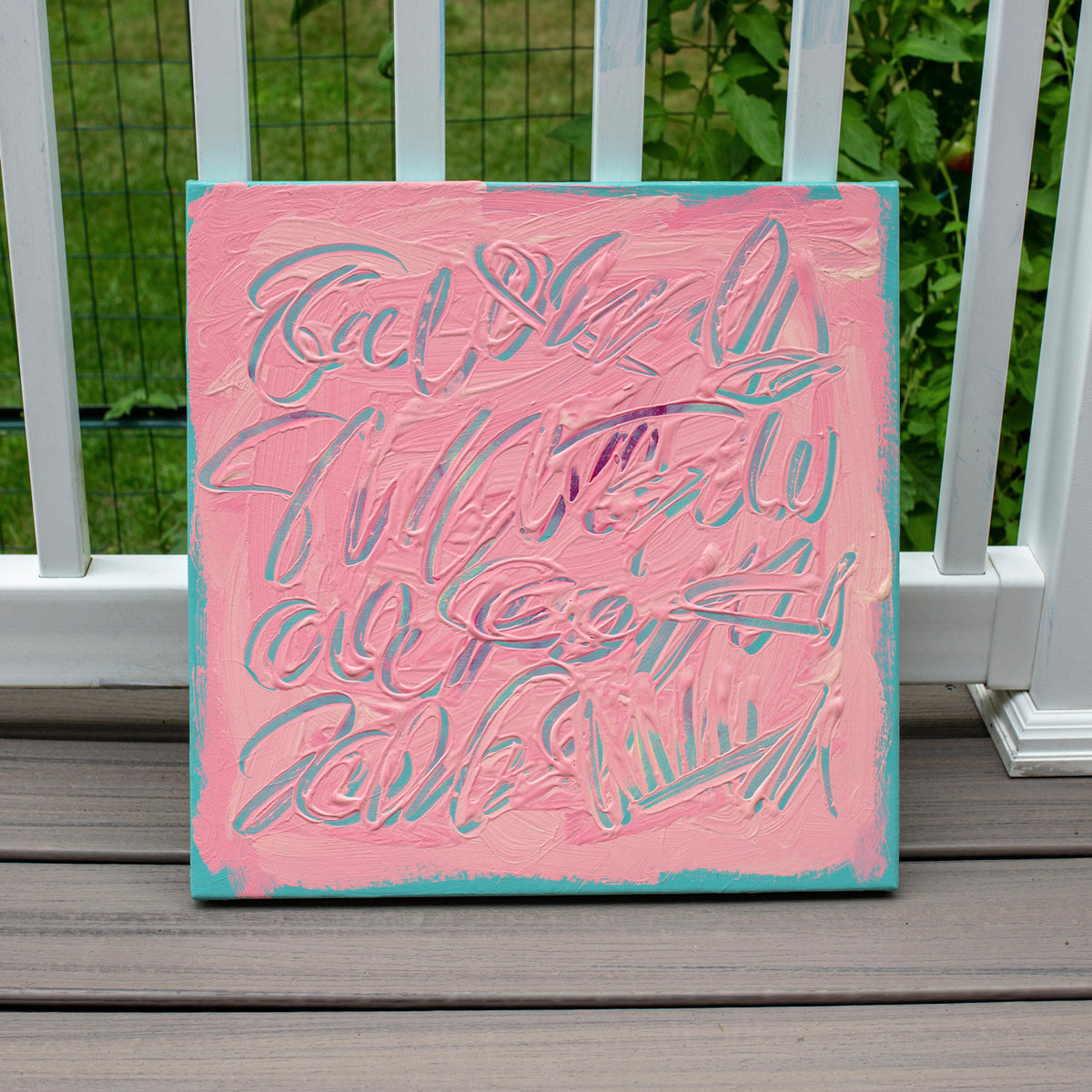 A square painting with a pink background and textured abstract lettering, leaning against a white railing on a wooden deck with green foliage in the background.