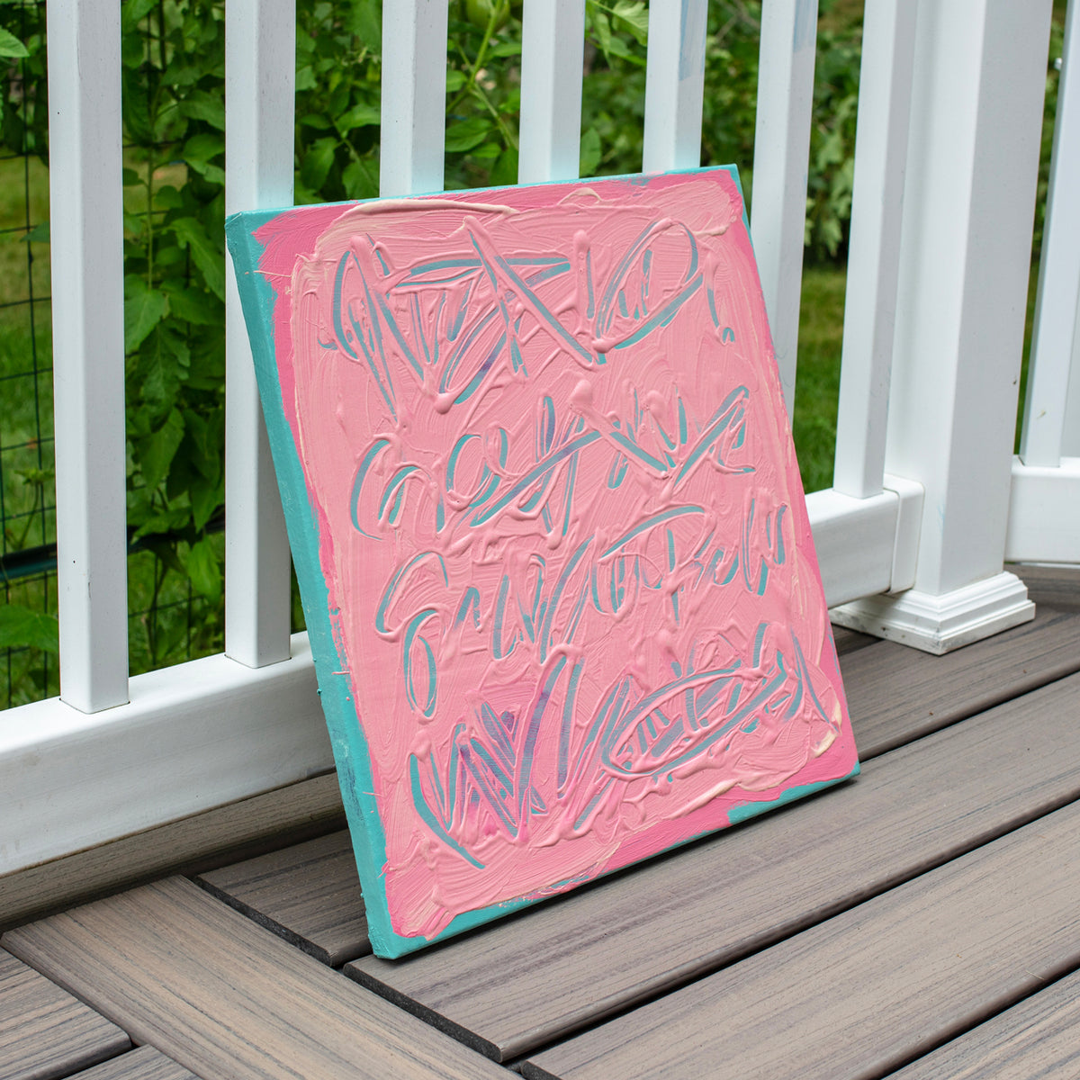 A square canvas art print with thick pink and teal paint textures and abstract lettering, leaning against a white wooden deck railing with green foliage in the background.