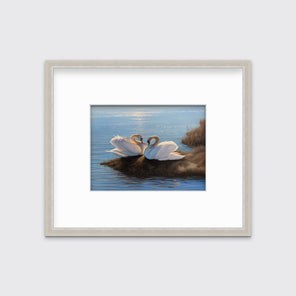 A print of two swans facing each other on an embankment next to water matted in a silver frame hangs on a white wall.