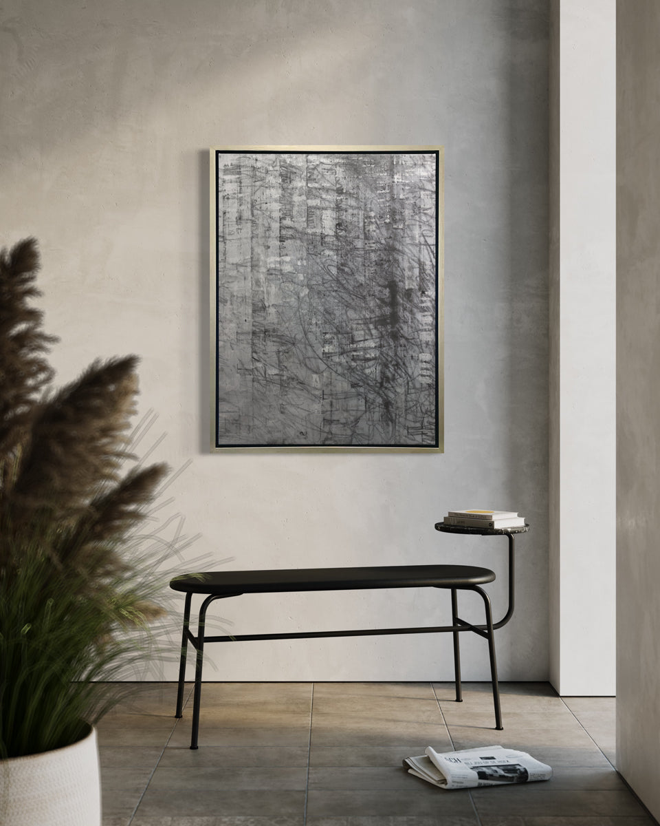 An abstract silver-grey limited edition art print by Ned Martin in a silver frame hangs on a white wall in a room with a black bench in front of it.