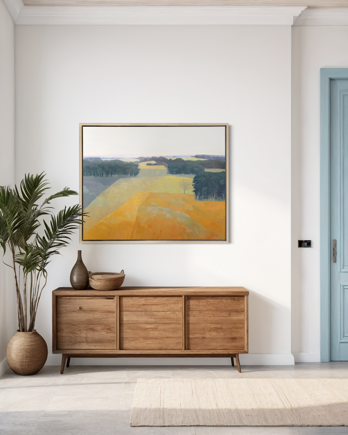 A light blue, green and light orange abstract landscape print in a white oak wood floater frame hangs on a white wall above a low natural wood credenza with a vase, an open basket and a large potted floor plant.