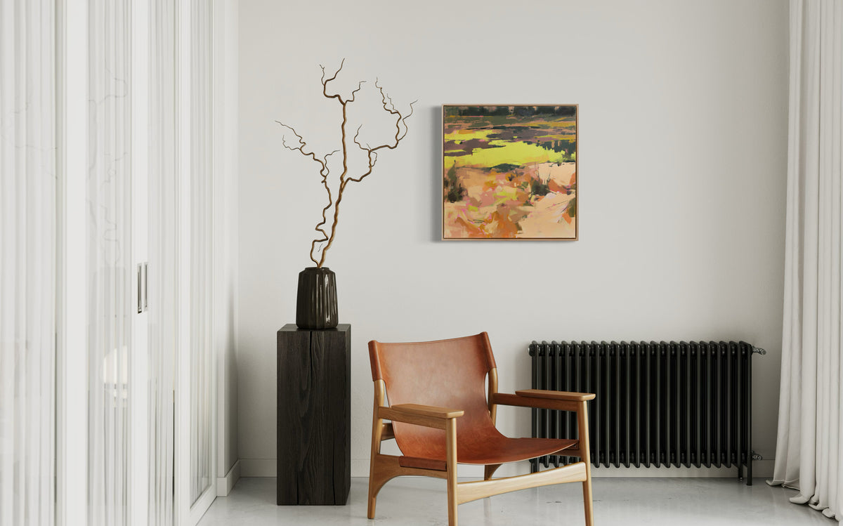 A yellow and orange abstract landscape painting hangs on a white wall over a black radiator. A chair and a black wooden pedestal with with a black vase holding decorative twigs sits in front.