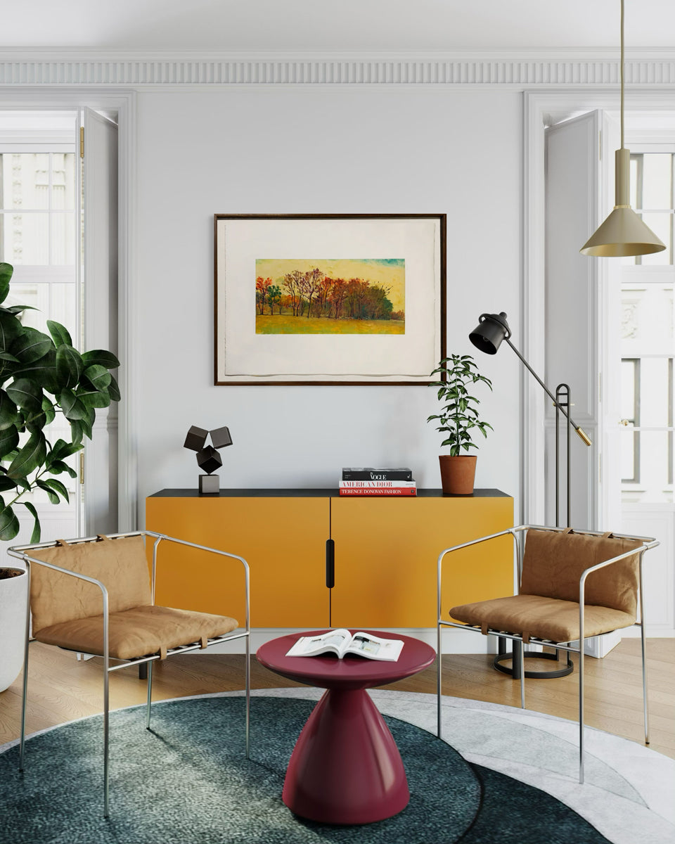 Framed painting of a landscape with trees and a yellow sky on a white wall hanging over an orange credenza.