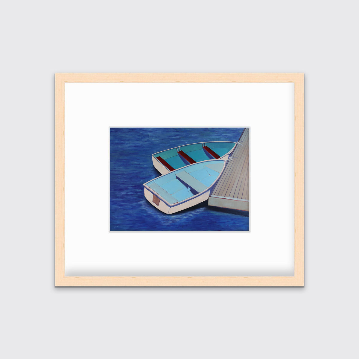 A blue, light blue and beige contemporary nautical print of two docked rowboats in a natural wood frame with a mat hangs on a white wall.
