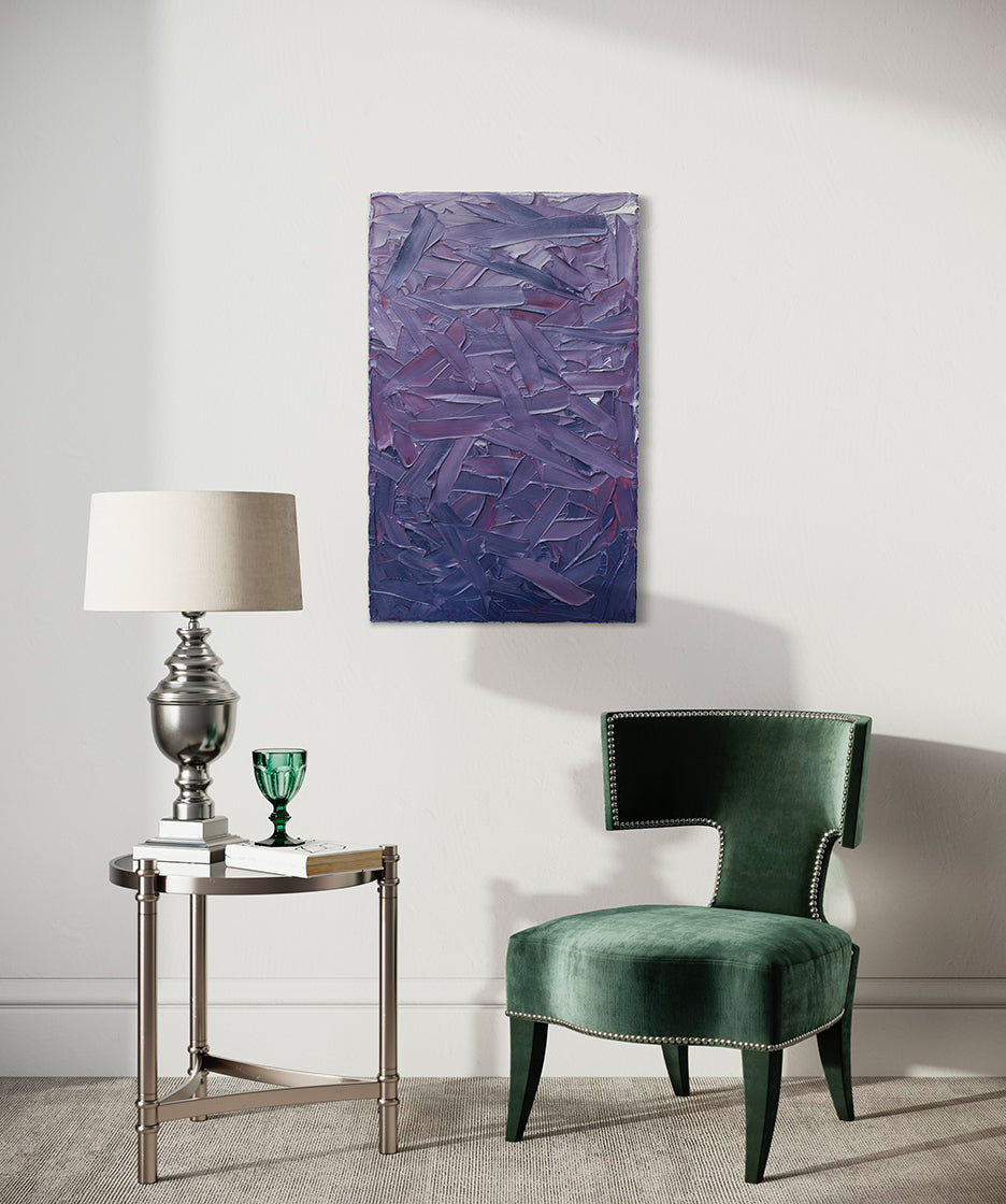 A tonal purple and pink textured abstract painting by Teodora Guererra hangs on a white wall above a green velvet chair and an end table of glass and crome with books and a table lamp of silver with a cream shade. On one of the books sits a green glass.