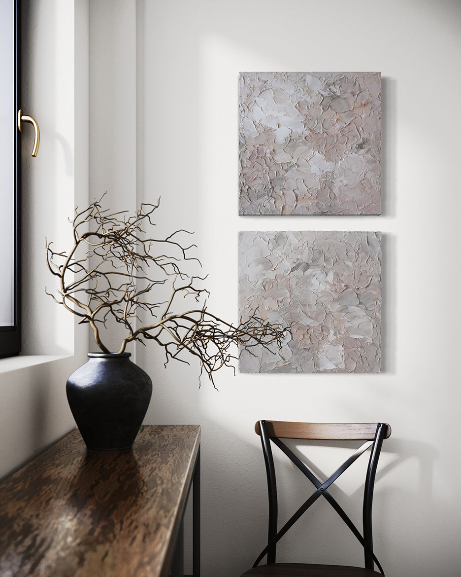 A pair of thickly textured abstract paintings in light pink, grey, coral and white by Teodora Guererra hanging on a white wall over a chair. To the left is a wood bench with a black ceramic vase holding tree branches in it. Beyond is a window.