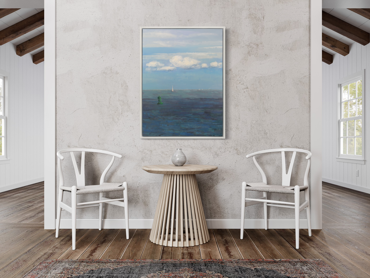 A coastal seascape print in a white floater frame hangs on a white wall above a light wood pedestal table with a vase and two white white chairs.