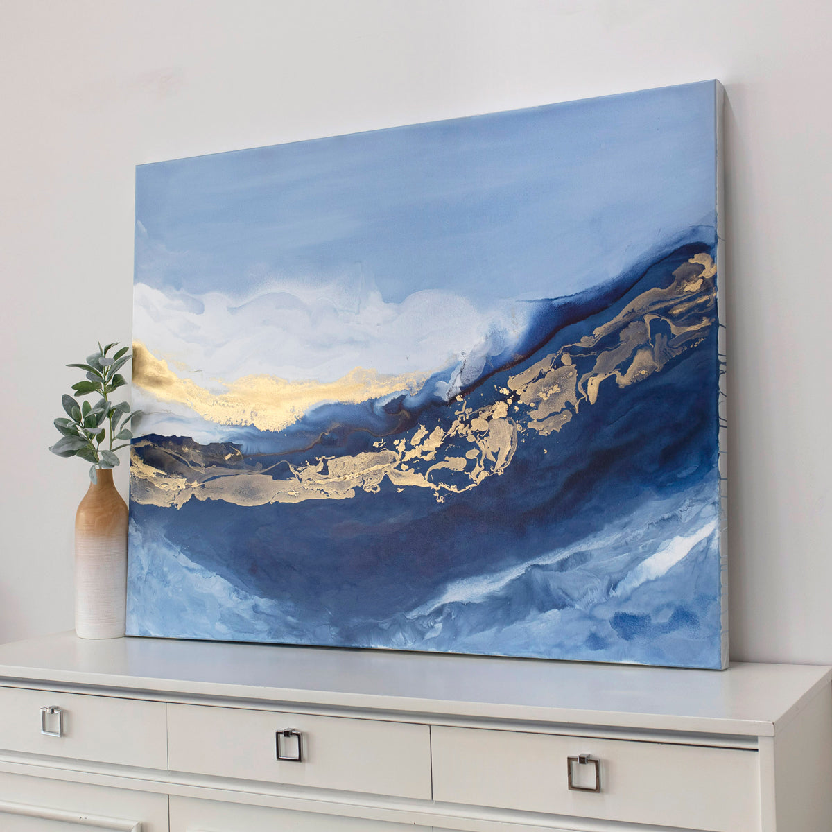 Abstract painting with blue, gold, and white on a wall above a white cabinet.
