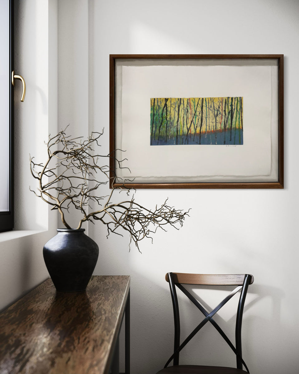Framed artwork of a forest scene with trees in yellow and green hanging on a white wall over a chair by a window.