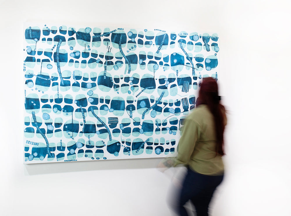 A woman in a green shirt walks in front of a blue and white abstract painting hanging on a white wall.