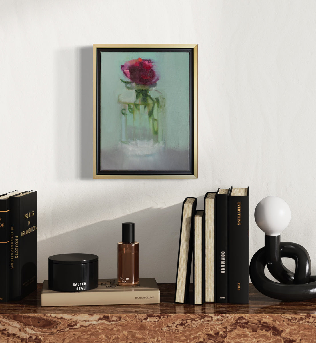 A small framed painting of a red flower in a clear glass vase, framed in a gold floating frame, hangs on a white wall above a red marble table surface with black and white table decor.