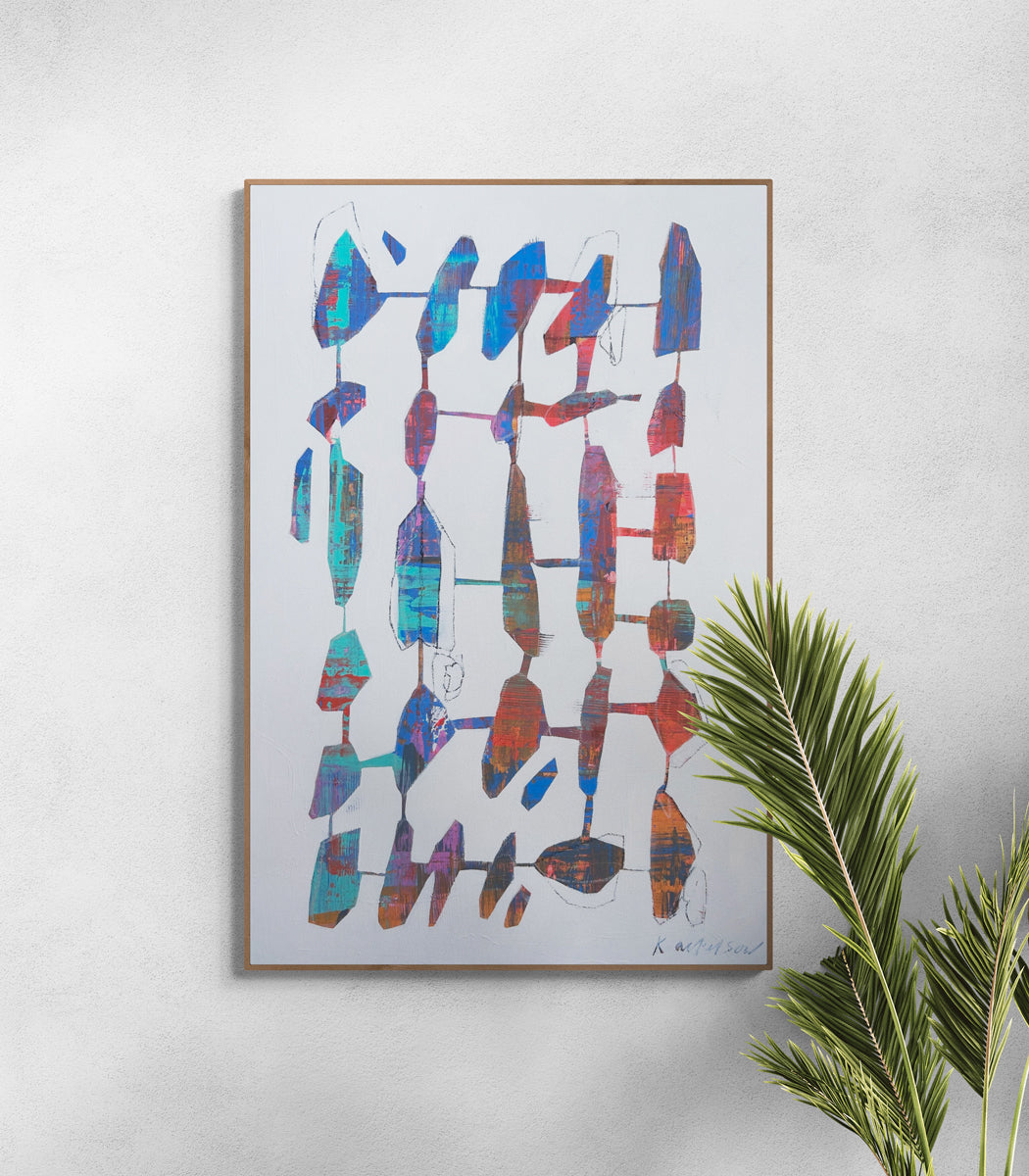 Framed abstract artwork with colorful geometric shapes on a white wall above a plant.